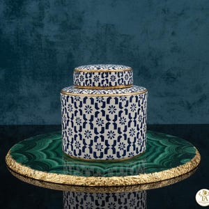 Puede incluir: Un tarro de cerámica decorativo con tapa, con un patrón floral azul y blanco y ribete dorado. El tarro se exhibe sobre una bandeja decorativa verde y dorada. El fondo es de color verde azulado oscuro.