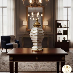 Könnte beinhalten: Eine weiße und goldene dekorative Vase mit Deckel steht auf einem dunklen Holztisch. Die Vase hat ein geometrisches Design mit goldenen Akzenten. Der Raum hat einen klassischen, eleganten Stil mit einem Kronleuchter und einem dunkelblauen Sessel.