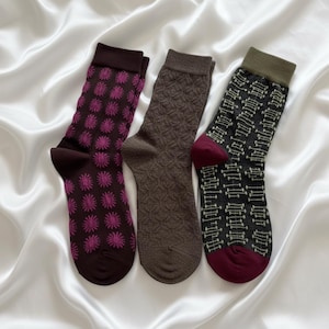 May include: Three pairs of socks in shades of brown, with unique patterns. One pair features a floral design in magenta, another has a textured pattern, and the third displays a geometric pattern in black and olive green with burgundy accents.