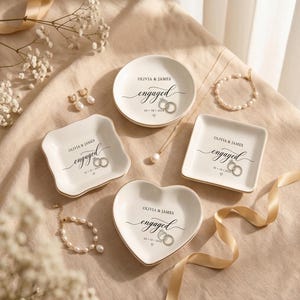 May include: A collection of white ceramic ring dishes in various shapes, including round, square, and heart-shaped. Each dish is inscribed with "Olivia & James engaged" and the date. Gold trim accents the edges. Jewelry and ribbons are also present.