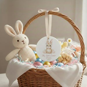 Puede incluir: Una cesta de Pascua de mimbre tejida llena de huevos coloridos, un conejito de peluche y golosinas. Un adorno redondo con una ilustración de conejito cuelga del asa. El adorno dice "My First Easter Emma 2026".