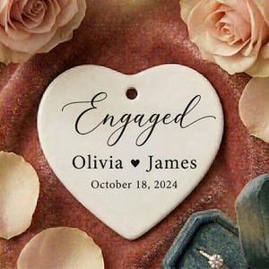 Peut inclure: Un souvenir de fiançailles en céramique blanche en forme de cœur avec le mot "Engaged" en écriture cursive. Il est inscrit "Olivia ♥ James" et "October 18, 2024". Entouré de roses roses et d'un écrin à bagues turquoise.