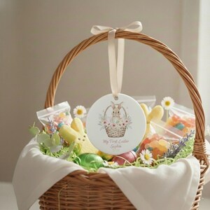 Puede incluir: Una cesta de Pascua tejida llena de golosinas, que incluye un adorno de cerámica con una ilustración de un conejito. La cesta contiene caramelos, huevos decorativos y un dulce de malvavisco con forma de conejito amarillo. Una cinta blanca está unida al adorno.