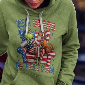 Può includere: Felpa con cappuccio verde con un personaggio simile al Grinch e un cane seduto in poltrona con un design patriottico. La grafica include il testo "250 YEARS OF FREEDOM" in rosso e blu, che celebra un evento storico.