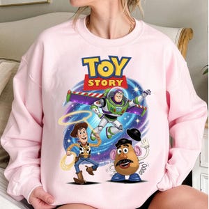Peut inclure: Un sweat-shirt rose clair avec l'inscription "TOY STORY" au-dessus d'un dessin de Buzz l'Éclair, Woody et M. Patate. Buzz l'Éclair est en vol, Woody tient un lasso et M. Patate fait signe.