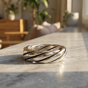 May include: A silver-colored ring with a twisted rope design. The ring is made of a shiny metal and sits on a marble surface. The ring's design features multiple parallel grooves that create a textured appearance.