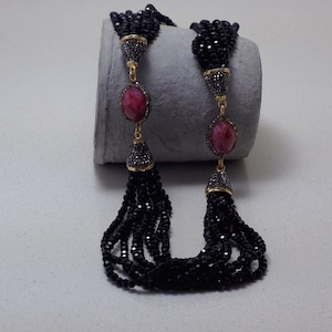 May include: A black beaded necklace with a tassel design. The necklace features a gold chain with a large oval-shaped pink gemstone. The tassels are made of multiple strands of small black beads.