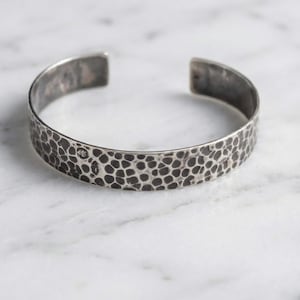 Puede incluir: Un brazalete de plata con textura martillada. El brazalete abierto presenta un patrón oscuro y moteado en su superficie. El metal tiene una apariencia ligeramente envejecida, lo que sugiere una pieza vintage o artesanal. El brazalete se muestra sobre una superficie de mármol.