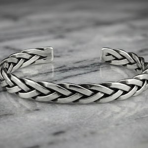 May include: A silver cuff bracelet with a braided design. The open-ended bracelet features interwoven strands, creating a textured, woven appearance. The bracelet is displayed on a marble surface.