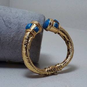 May include: A gold-colored bangle bracelet with a textured surface and turquoise-colored stone accents. The bracelet has a hammered texture and features turquoise stones at each end. The bracelet is open and has a hinge.