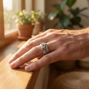 Puede incluir: Un anillo plateado con un diseño tejido se muestra en una mano. El intrincado patrón del anillo es el punto focal, con la luz reflejándose en el metal. El fondo incluye un alféizar de madera y plantas en macetas.