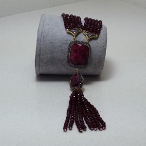 Op de afbeelding: Een opvallende ketting met een grote, vierkante, robijnrode edelsteenhanger en een druppelvormige steen eronder. Het heeft een kwast van kleine, bordeauxrode kralen en goudkleurige accenten.
