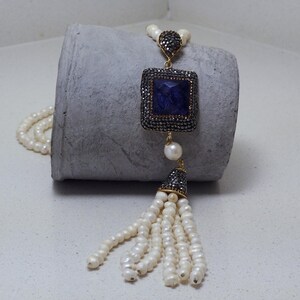 May include: A long necklace with a square, dark blue pendant and a pearl tassel. The pendant is framed by a silver-coloured border. The necklace also features a strand of small, white pearls.