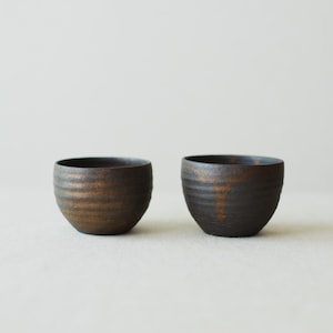 May include: Two small, round ceramic cups with a dark, textured finish. The exterior surfaces feature horizontal ridges and mottled brown and black tones, with a smooth, dark interior rim. The cups have a rounded base and a wide, open top.