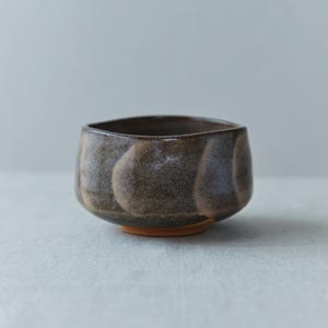 May include: A handmade ceramic bowl with a dark brown glaze and a textured surface. The bowl has a rounded shape with a flat base and a slightly flared rim. The base is a light orange color.