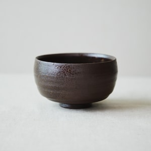 Può includere: Una ciotola in ceramica marrone scuro con una finitura opaca testurizzata e una superficie leggermente irregolare e rustica. La ciotola presenta una forma arrotondata con un sottile rilievo orizzontale vicino al bordo e una piccola base circolare.