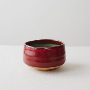 以下が含まれることがあります： 外側は光沢のある深い赤色の釉薬で覆われ、縁に向かって黒に近い色に変化する小さな丸い陶器のボウル。ボウルの内側は落ち着いた淡い緑色の釉薬が施されており、底面は釉薬のかかっていない明るい茶色の粘土のままとなっている。