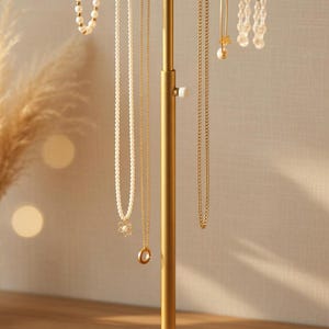May include: A gold-colored jewelry display stand with a marble base. The stand holds various necklaces, bracelets, and earrings. The jewelry includes pearl strands, delicate chains, and beaded earrings. The stand is on a wooden surface.