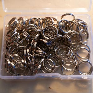 May include: A clear plastic container filled with numerous silver-colored split key rings. The key rings are tightly packed together, with many visible. The container is open, and the key rings are ready for use.