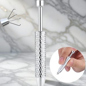 Puede incluir: Una herramienta plateada con un mango con diseño de diamantes y una pequeña parte superior redondeada. La herramienta tiene un eje largo y delgado y un extremo en forma de garra. Primer plano.