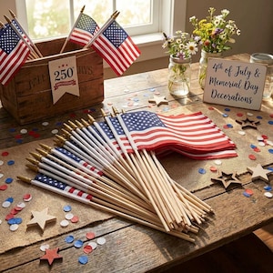 May include: A collection of American flags with wooden poles, displayed on a rustic wooden table. The flags are red, white, and blue, with stars and stripes. The scene includes a wooden crate, wooden stars, and a sign that reads "4th of July & Memorial Day Decorations."