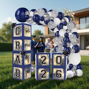 May include: A graduation party decoration set featuring navy blue, silver, and white balloons forming an arch. Illuminated boxes spell out "GRAD" and "2026", with a graduation cap icon. A family celebrates in the background.