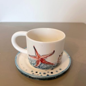 Op de afbeelding: Witte keramische koffiekop en schotel set. De beker heeft een handgeschilderd rood zeester ontwerp met blauwe accenten. De schotel heeft een blauwe rand en decoratieve blauwe stippen. Een charmant nautisch thema.