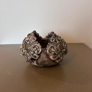 May include: A small, decorative ceramic bowl with ornate, raised floral designs. The bowl features a mix of colors, including pink, green, and brown, with a textured surface. The floral accents are highlighted with gold-colored details.