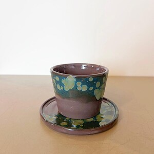 Può includere: Un set di tazza e piattino in ceramica. La tazza è di colore marrone scuro con un motivo maculato verde e blu acqua sul bordo. Il piattino rispecchia il design della tazza. Il set è appoggiato su una superficie marrone chiaro.