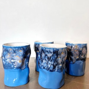 May include: Four ceramic tumblers with a unique design. The bottom half of each tumbler is a solid light blue, while the top half features a textured, dark blue glaze with gold accents. The interiors are white.