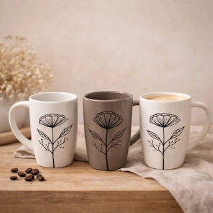 Puede incluir: Tres tazas de cerámica con un diseño floral. Una taza es blanca, otra es color topo y otra es blanca con café. Cada taza presenta un dibujo lineal negro de una flor y un tallo. Las tazas tienen asas blancas y están sobre una superficie de madera.