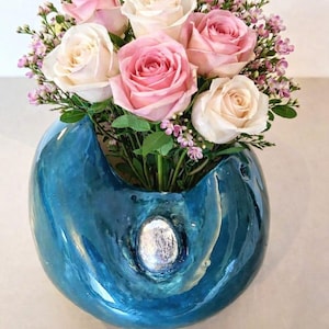 Könnte beinhalten: Ein lebendiges Blumenarrangement mit rosa und cremefarbenen Rosen, akzentuiert mit kleinen lila Blüten und grünem Laub. Die Blumen befinden sich in einer glänzenden, blauen Keramikvase mit silbernem Akzent.