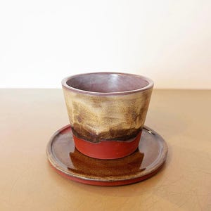 Può includere: Un set di tazza e piattino in ceramica fatto a mano. La tazza ha un esterno marrone e beige con una base rossa e un interno viola. Il piattino è marrone con un bordo rosso. Il set è progettato per servire bevande.