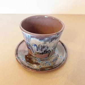 Pode incluir: Um conjunto de chávena e pires de cerâmica artesanal. A chávena é castanha escura com um esmalte azul e branco na borda. O pires é da mesma cor castanha com detalhes azuis. Perfeito para café ou chá.