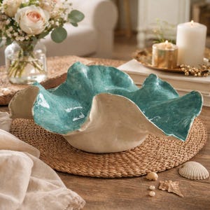 Può includere: Una ciotola decorativa in ceramica con un design ondulato unico. Il bordo è turchese e l'interno color crema. È posizionata su un sottobicchiere intrecciato, con conchiglie e perle. Un elemento decorativo per la casa.