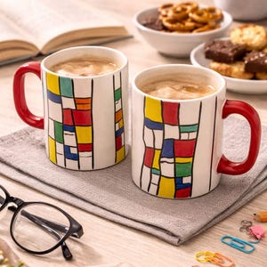 Könnte beinhalten: Zwei weiße Keramikbecher mit roten Griffen, gefüllt mit Kaffee. Die Becher haben ein farbenfrohes geometrisches Design in Rot, Gelb, Blau und Grün. Die Becher stehen auf einer grauen Serviette, mit Snacks und einem Buch im Hintergrund.
