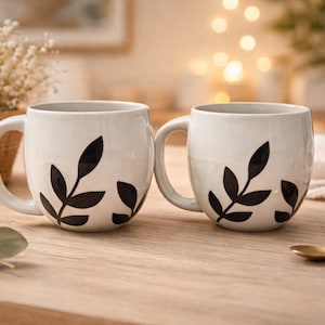 Può includere: Due tazze in ceramica bianca con disegni di foglie nere. Le tazze hanno grandi manici e sono appoggiate su una superficie di legno chiaro. Sono visibili anche un cucchiaino dorato e un cestino di fiori secchi.