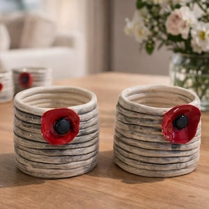 May include: Two ceramic candle holders with a textured, striped design. Each holder features a red poppy flower with a black center. The holders are cylindrical and sit on a wooden surface. A vase of flowers is in the background.