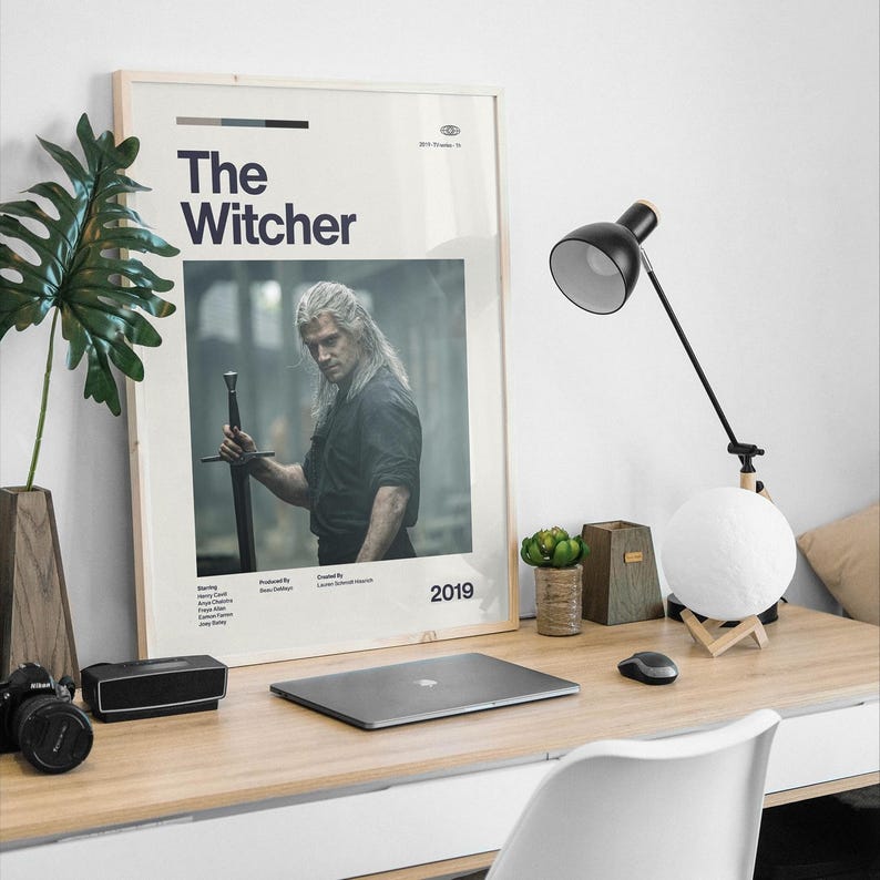 May include: A vertical rectangular poster featuring The Witcher with a central photograph of a person with long hair holding a sword. The poster includes the title, the year 2019, and production credits in black text on a white background, framed in light wood.