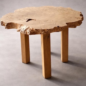 May include: A natural wood side table with a unique, organic shape. The tabletop has a live edge and is supported by three sturdy, square legs. The light brown wood grain is visible throughout the table.