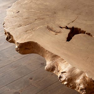 Puede incluir: Primer plano de una mesa de losa de madera natural con borde vivo. La mesa es de color marrón claro con vetas de madera visibles y una gran grieta. La mesa está sobre un suelo de madera.