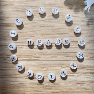 Peut inclure: Des perles blanches et rondes avec des lettres dorées épellent "BEADS" disposées en cercle. Des perles individuelles avec des lettres sont également disposées autour du cercle. Les perles sont sur une surface en bois brun clair.