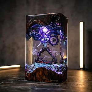 Puede incluir: Una pieza de arte de resina rectangular con una figura de personaje con un atuendo con capucha púrpura y blanco. La figura está rodeada de elementos arremolinados púrpuras y negros, encerrada en un bloque de resina transparente.
