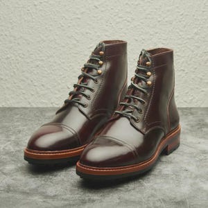 May include: A pair of burgundy leather boots with a cap toe design. The boots have black laces, brass eyelets, and a brown and black sole. The boots are approximately 6 inches tall.