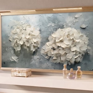 May include: A framed floral painting with a gold-coloured frame. The artwork features two large, textured white flower clusters against a light blue background. A white shelf below displays a decorative box and several glass bottles.