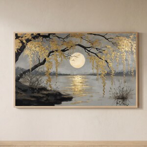 May include: A framed landscape painting depicts a moonlit scene over a lake. The artwork features a large, bright moon, golden-leafed branches, and a reflective body of water. The color palette includes shades of gray, gold, and black.