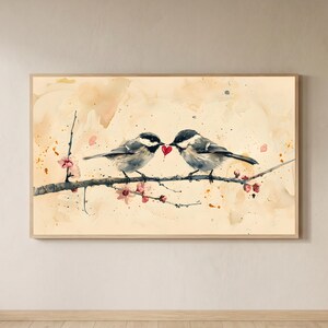 Puede incluir: Pintura a la acuarela de dos pájaros posados en una rama, uno frente al otro con un corazón rojo entre sus picos. Los pájaros tienen plumas grises y negras. Flores rosas y un fondo beige claro completan la obra de arte, enmarcada en madera clara.