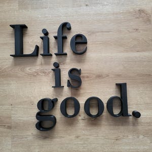 Pode incluir: Letras de madeira pretas que soletram "Life is good" em uma superfície de madeira marrom clara. As letras estão em uma fonte sem serifa e são espaçadas uniformemente. O ponto no final de "good" também é preto.