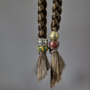 May include: Two brown braids with beads. The braids are tied with a silver bead, a green and yellow bead, and a gold bead. The other braid is tied with a gold bead and a red bead.