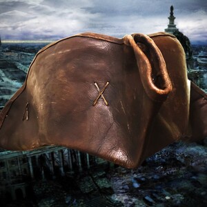 May include: A brown leather pirate hat with a stitched X on the front. The hat is worn and has a distressed look.
